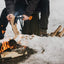 Persona che spacca legna con un'ascia Fiskars vicino a un fuoco da campo su neve, attrezzatura da campeggio invernale, abbigliamento outdoor, survivalismo, escursionismo invernale, preparazione del fuoco, attrezzi da campeggio, avventura nella natura.